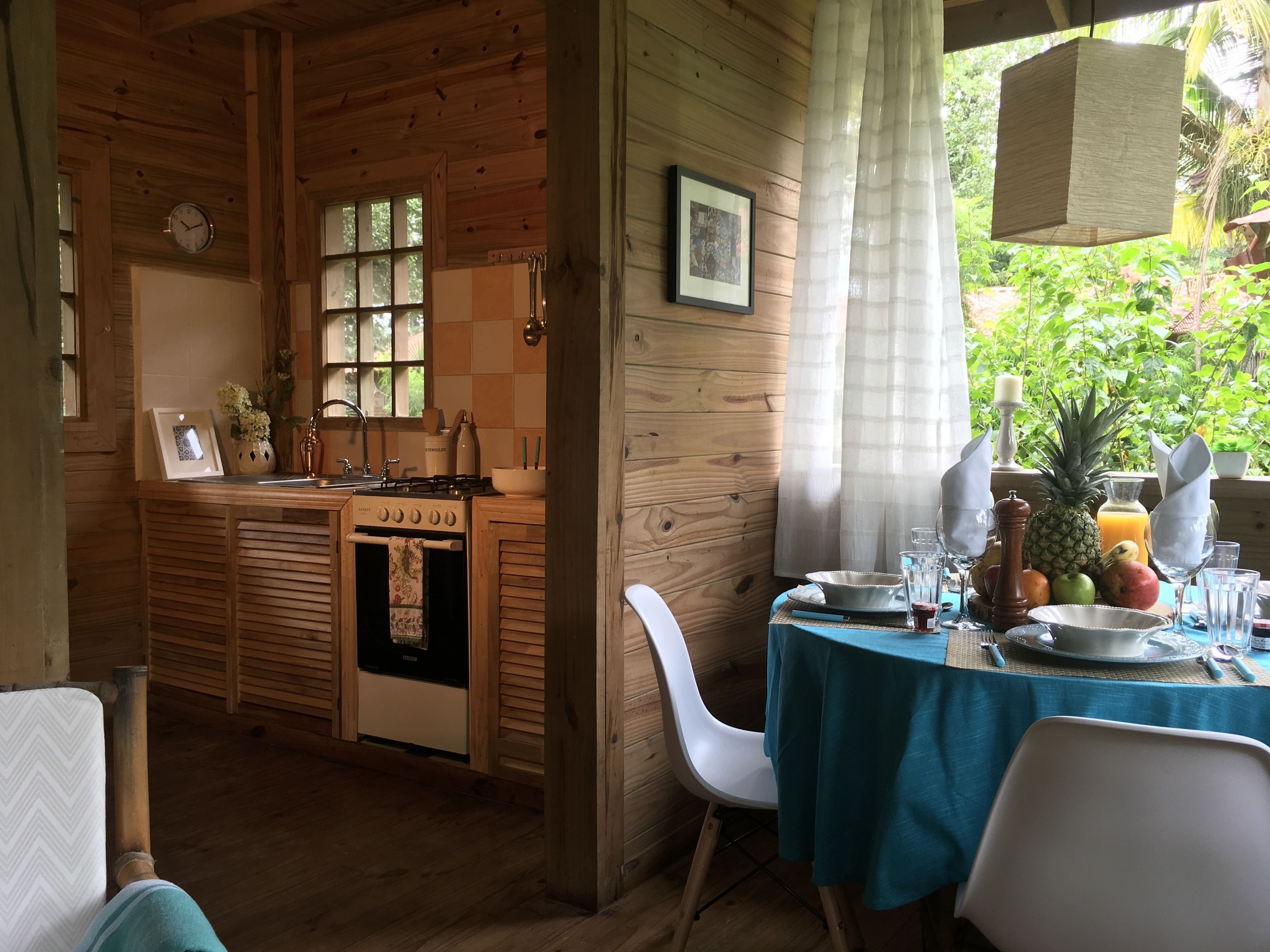 Une cabane en bois avec un coin cuisine, une table à manger dressée avec des assiettes, des verres et un panier de fruits.