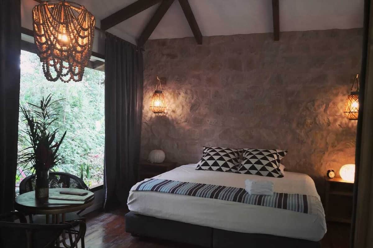 A bedroom with a large bed, a dining table with chairs, a chandelier, and a stone wall.
