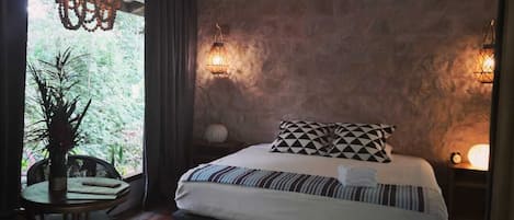A bedroom with a large bed, a dining table with chairs, a chandelier, and a stone wall.