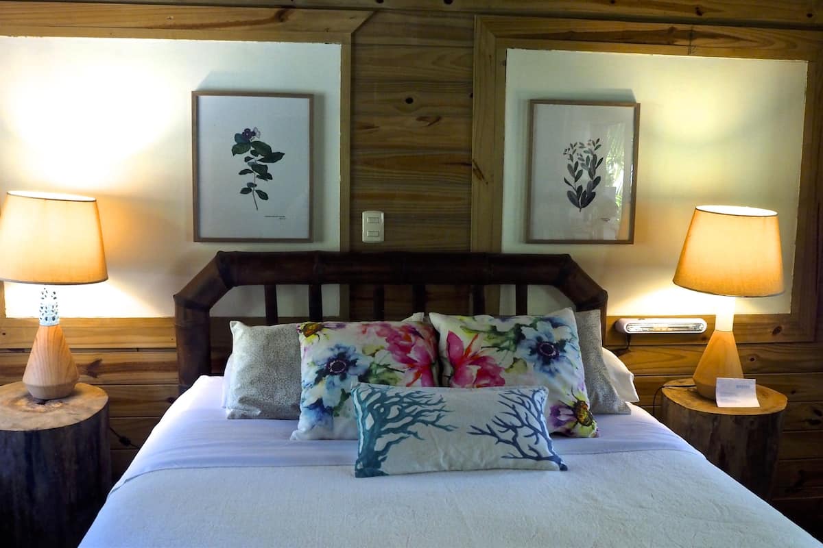 A bed with floral pillows, two wall-mounted lamps, and framed botanical prints on the wall.