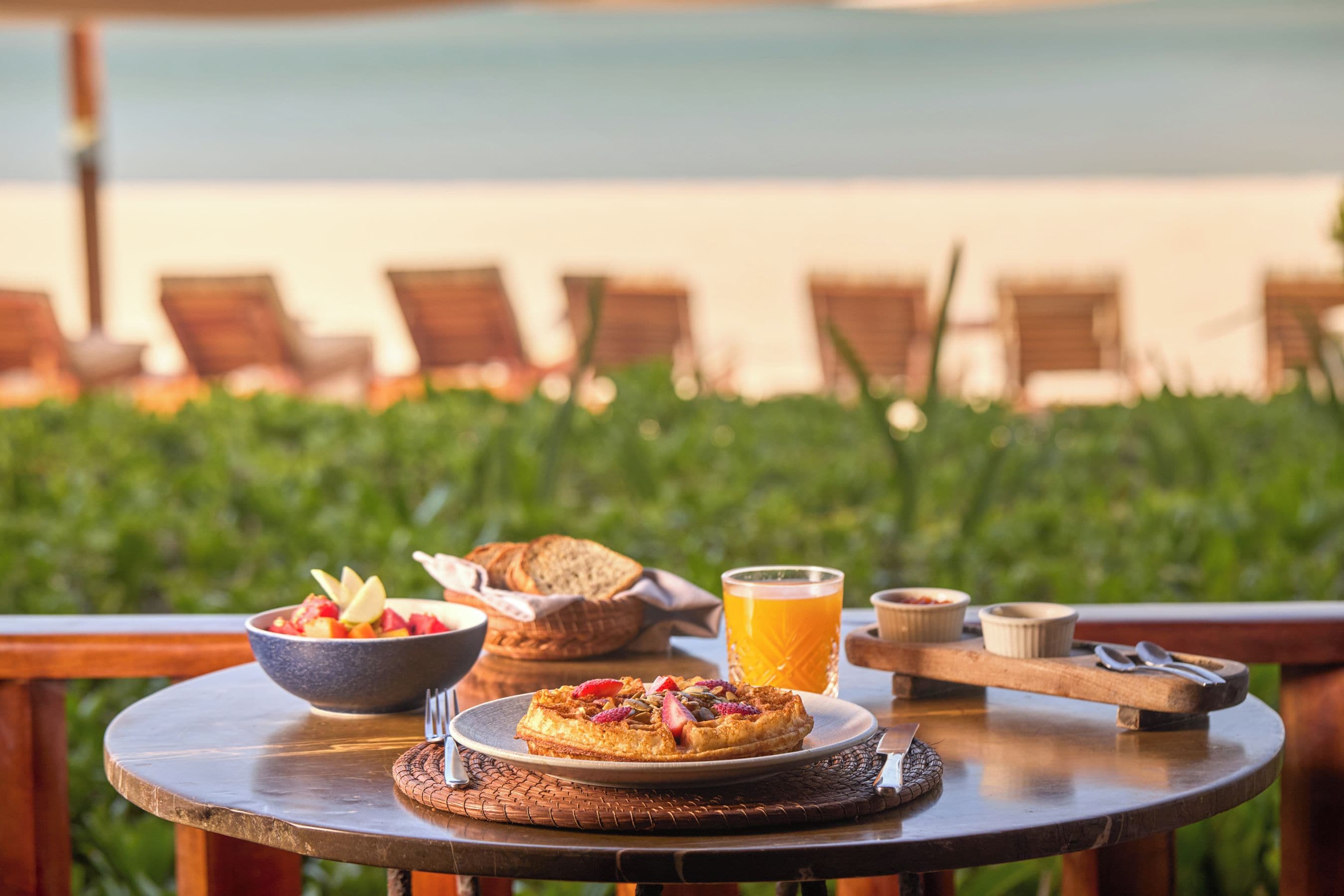 Una mesa con alimentos para el desayuno, incluyendo un tazón de fruta, una fuente de waffles, un vaso de jugo de naranja y una cesta de pan.