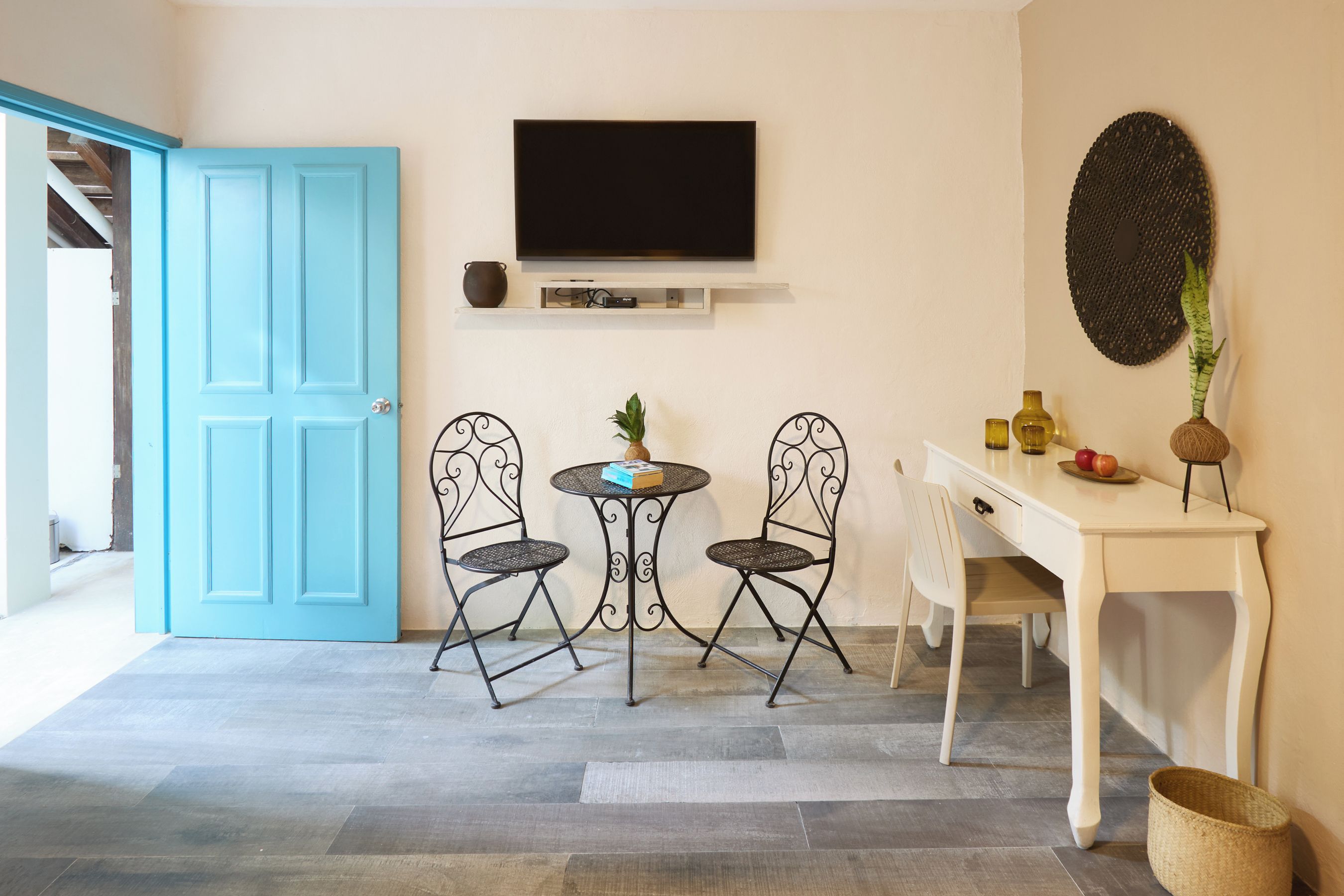 A room with a blue door, a flat-screen TV mounted on the wall, a small round table with two chairs, a white desk, and a woven wall hanging.