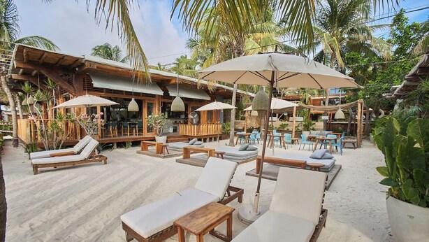 A beachside resort with wooden lounge chairs and umbrellas, surrounded by palm trees and a wooden building with outdoor seating. - Hotel Siesta Holbox (Isla Holbox)