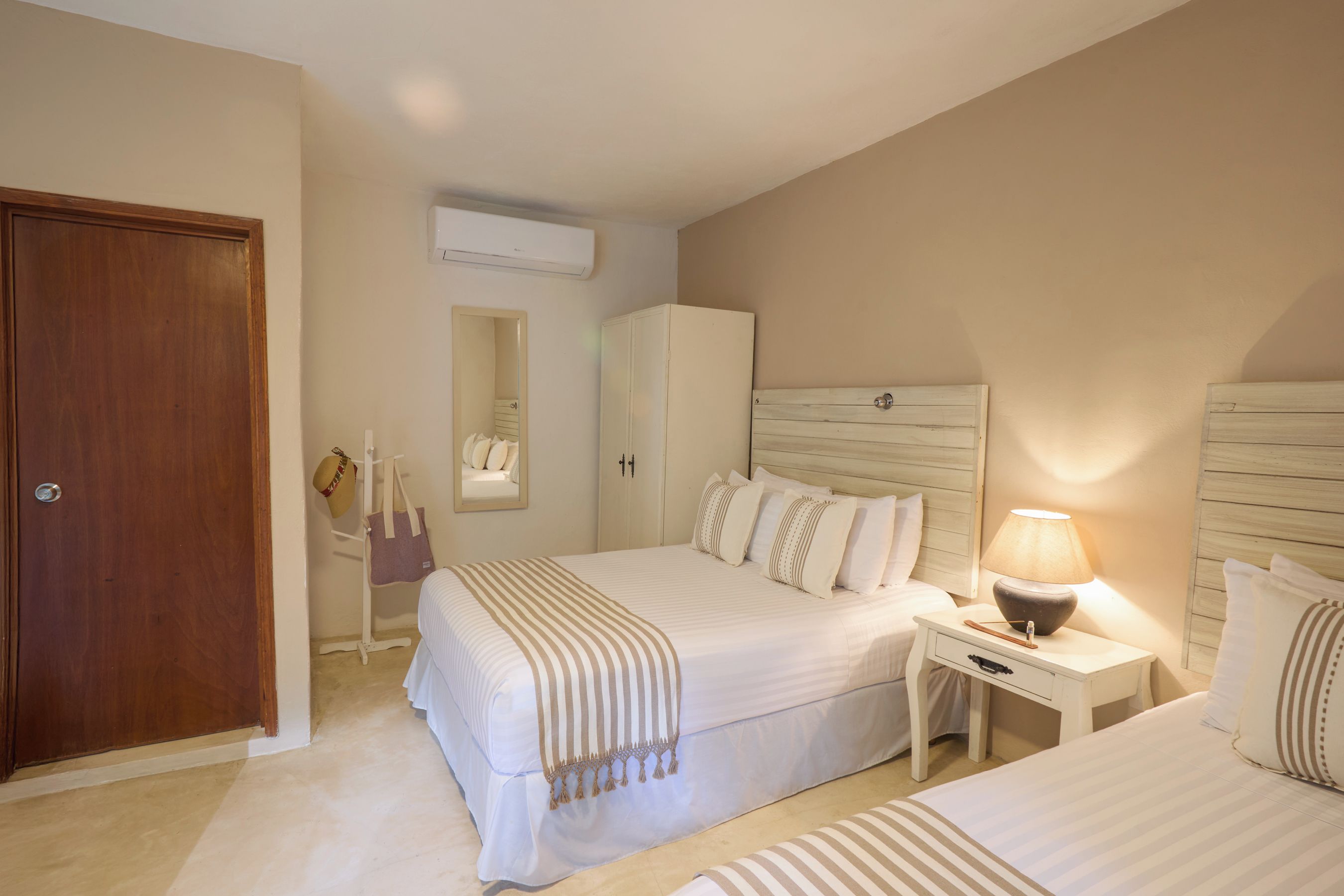 Une chambre d’hôtel avec une porte en bois, une armoire, un lit avec une literie à rayures, une table de chevet avec une lampe et un appareil de climatisation.