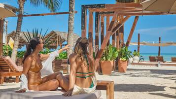 Dos personas sentadas en una tumbona en una playa arenosa con palmeras y una estructura de madera al fondo.