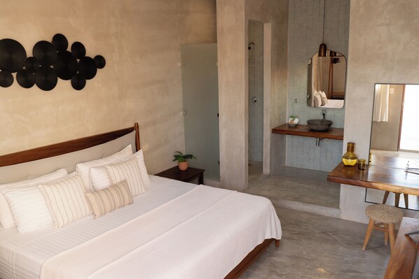 Un dormitorio con una cama grande, una mesita de noche con una planta, un baño con espejo y lavamanos, y un taburete de madera.