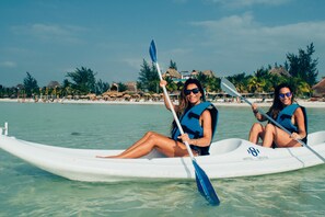 Dos personas en un kayak blanco en una playa con cabañas de techo de paja y palmeras al fondo.
