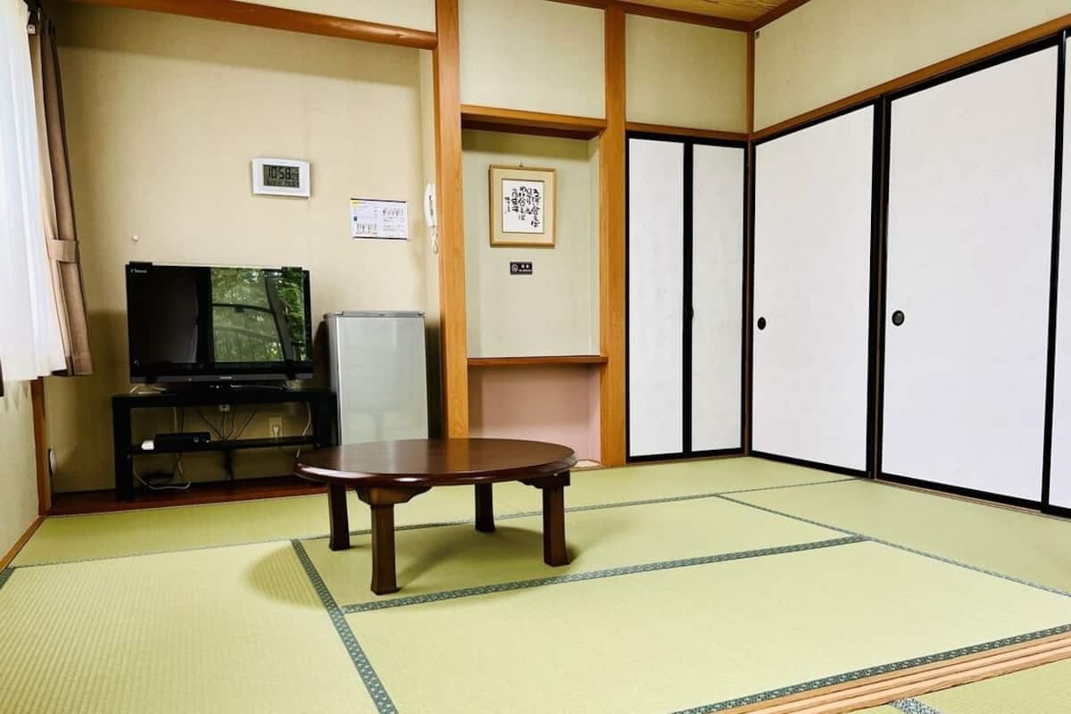 Ein Raum mit Tatami-Matten, einem Fernseher, einem kleinen Tisch und verschiebbaren Türen.