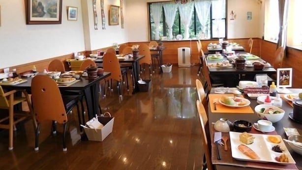 A dining area with wooden tables and chairs, a large window with a view of trees, and various dishes and condiments on the tables. - Pension AJIWAI (Nasu)