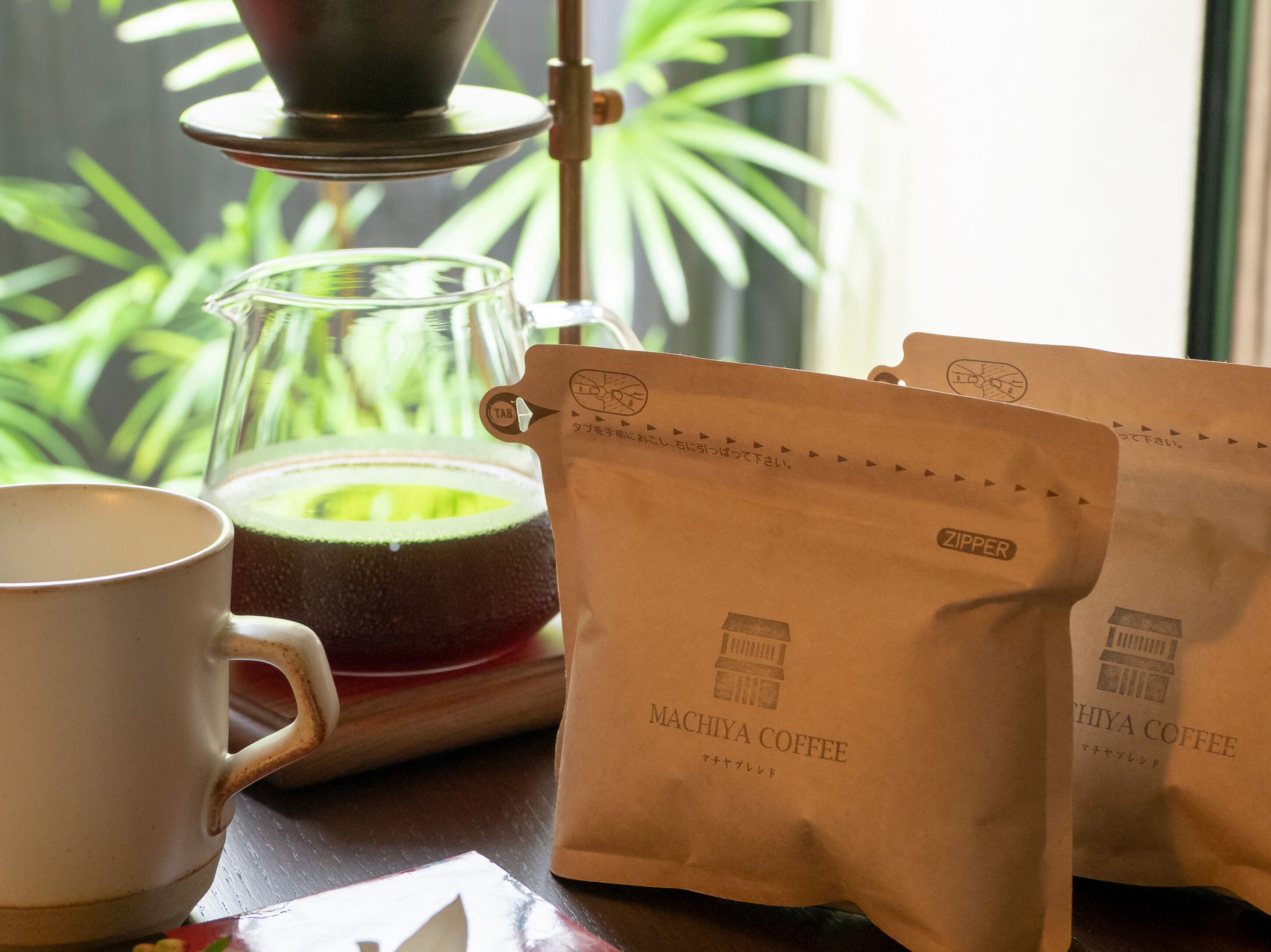 A coffee setup with a coffee maker, a glass carafe, and two bags labeled 'MACHIYA COFFEE'.