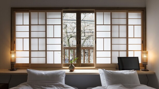 A hotel room with a large window, a bed with white bedding, a small table with a vase, and two lamps. - Hanagoromo (Kyoto)