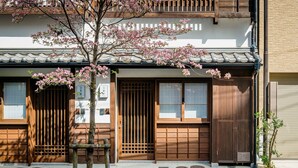 A traditional Japanese building with wooden sliding doors, a tiled roof, and a cherry blossom tree in front. - Hanagoromo (Kyoto)