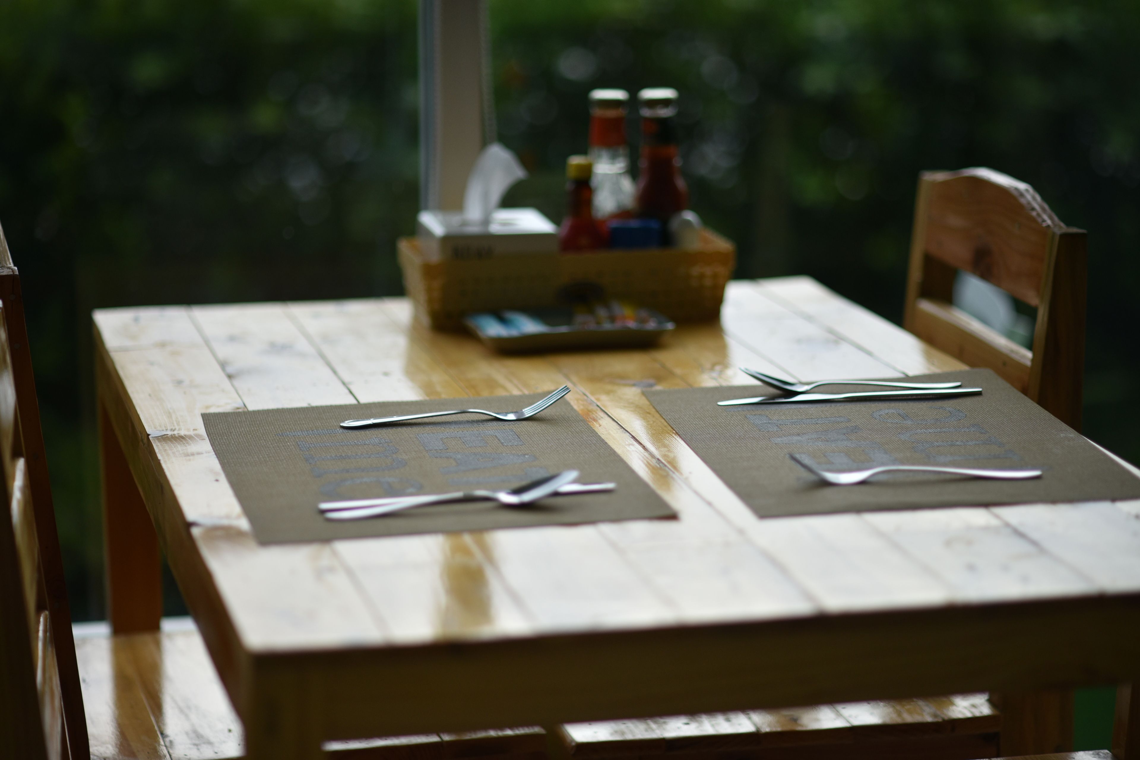 A wooden table with a placemat, cutlery, and condiments.