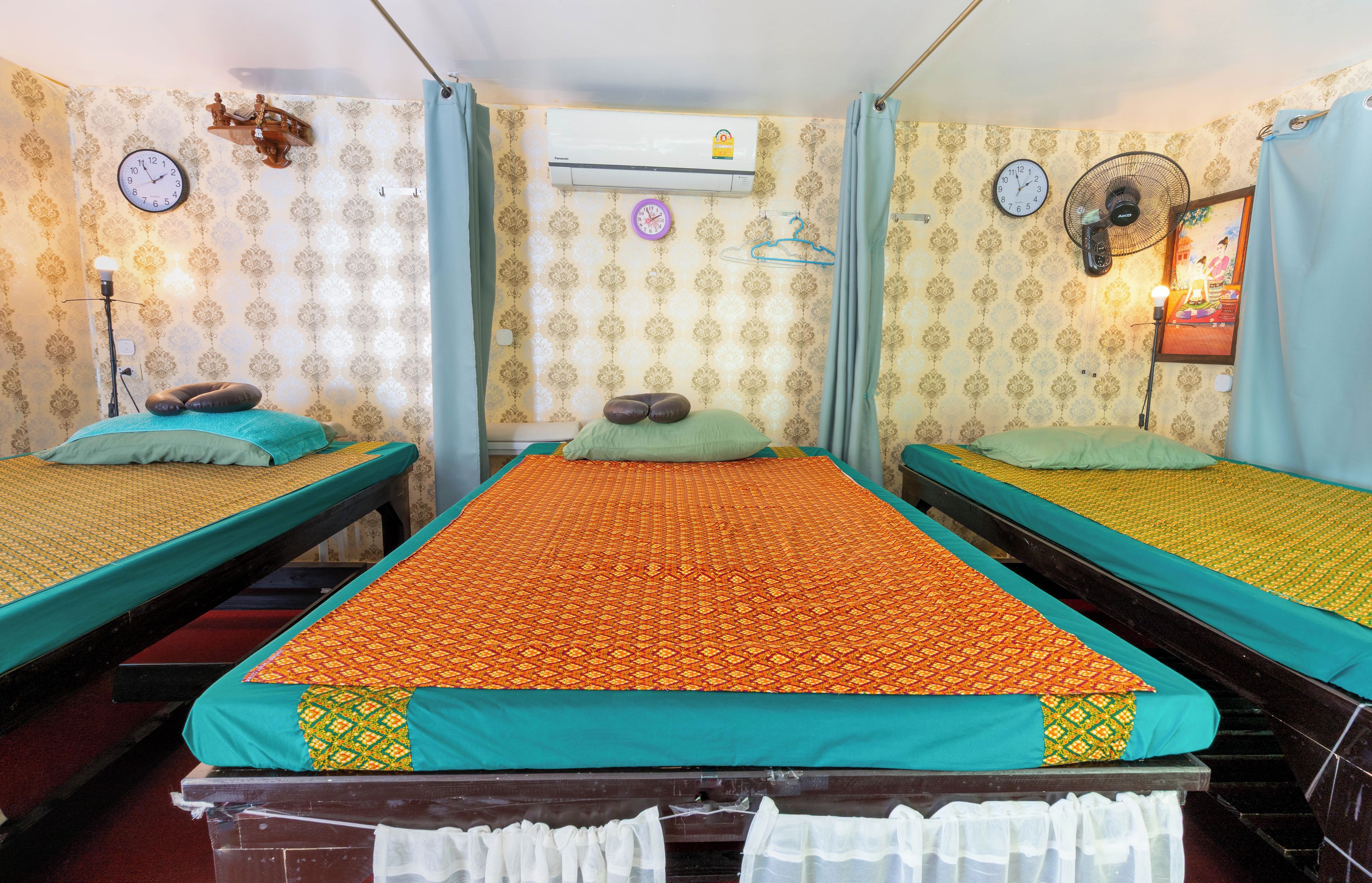 A room with three beds, each with a patterned bedspread, a wall-mounted clock, an air conditioning unit, and a wall-mounted fan.