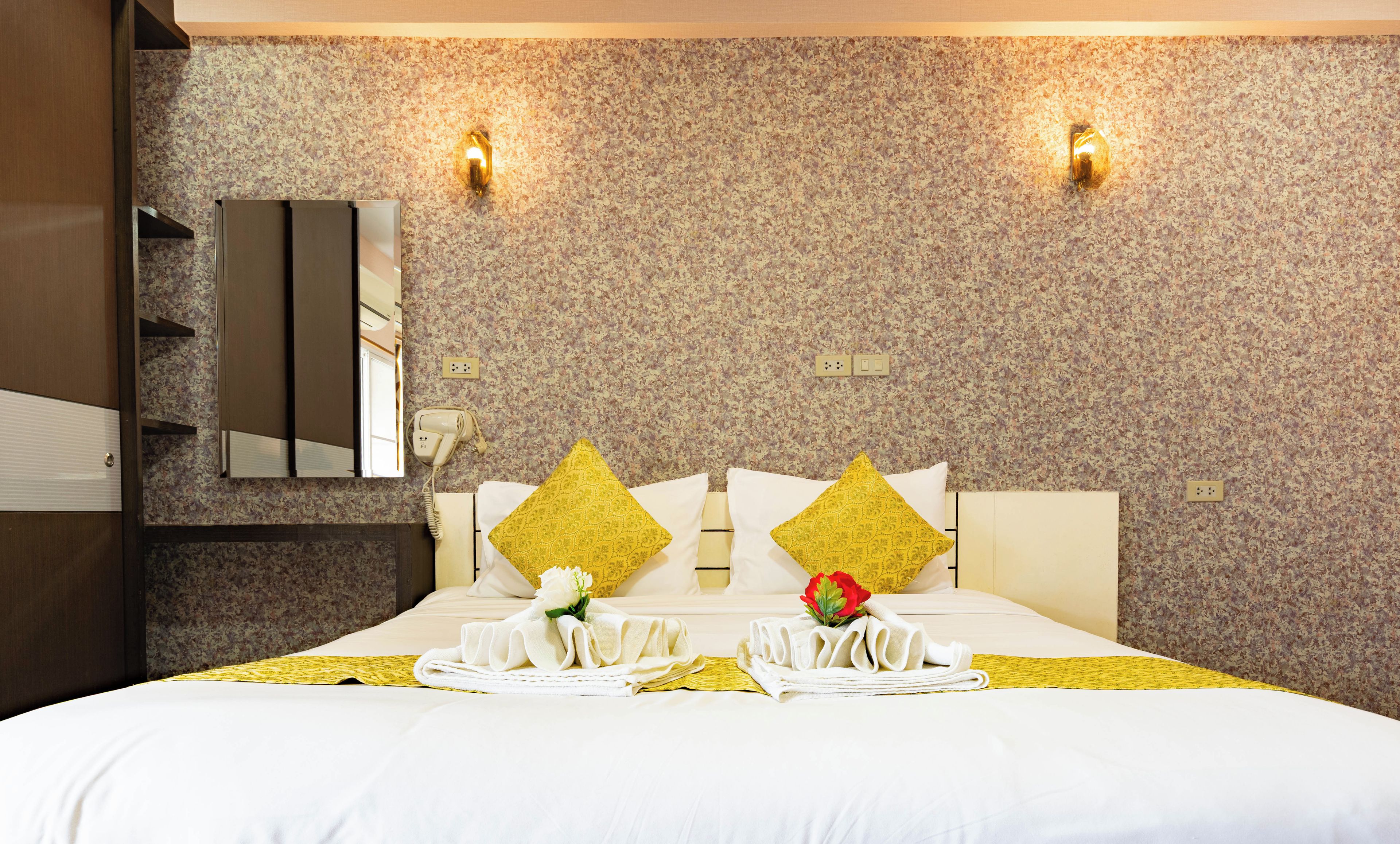 A hotel room with a large bed, two wall-mounted light fixtures, a mirror, a hairdryer, and a floral-patterned wall.