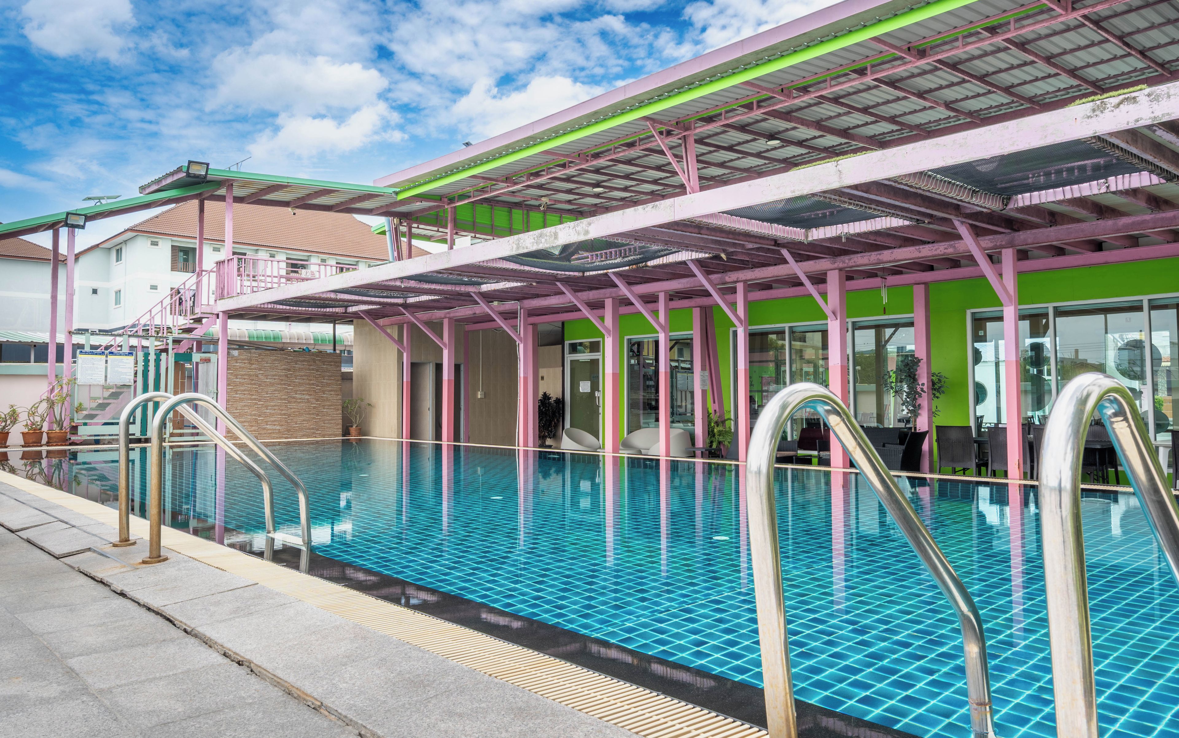 Ein Schwimmbecken mit überdachtem Bereich, Treppen und einem Gebäude mit grüner und pinkfarbener Fassade.
