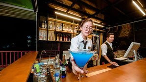 Zona de bar con estantes llenos de botellas de licor, un bartender preparando un cóctel azul y un monitor de computadora sobre la barra.