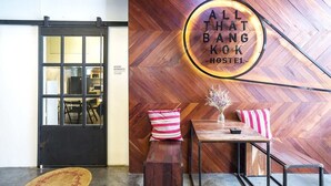 L'ingresso di un ostello con un pavimento in legno a spina di pesce, un cartello con la scritta "ALL THAT BANG KOK HOSTEL" e una panca con cuscini a righe.