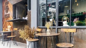 Area esterna con tavoli e sedie rotonde, una finestra con un cartello che indica la disponibilità di caffè freddo e vino, e un'arredamento rustico con piante essiccate.