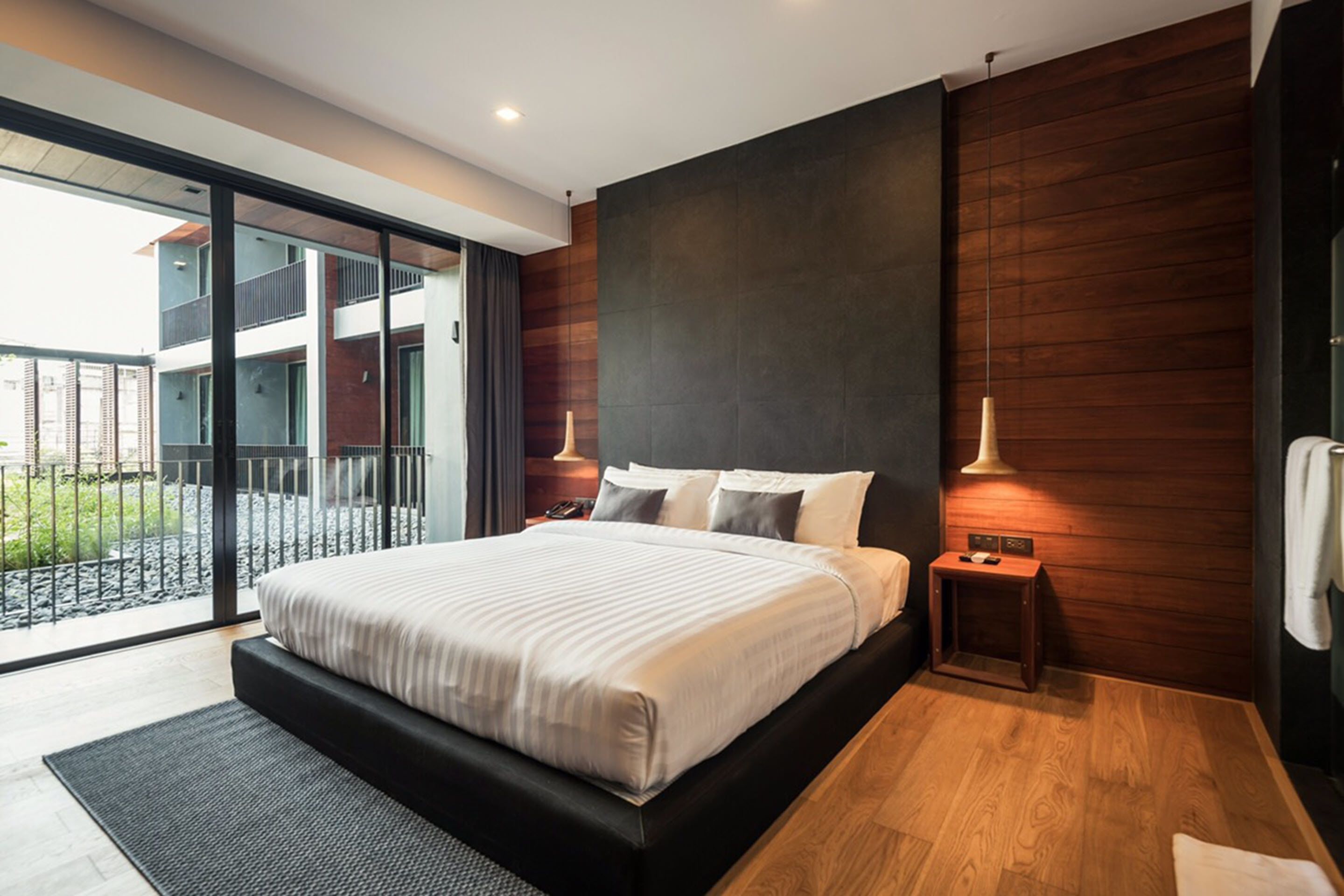 A modern bedroom with a large bed, wooden panel walls, a small bedside table, and a view of a balcony.