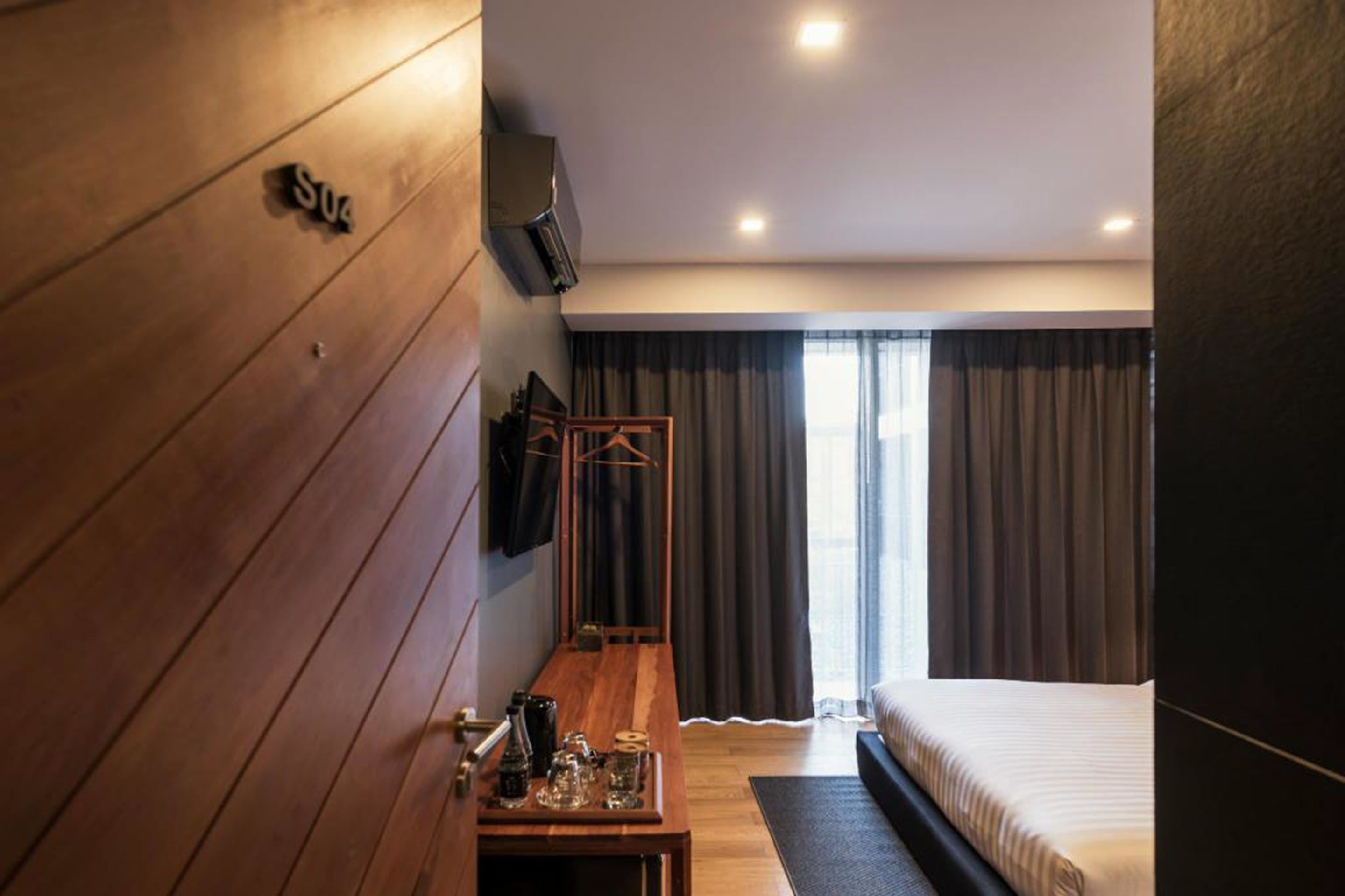 A modern hotel room with a wooden floor, a bed, a TV mounted on the wall, and a window with curtains.