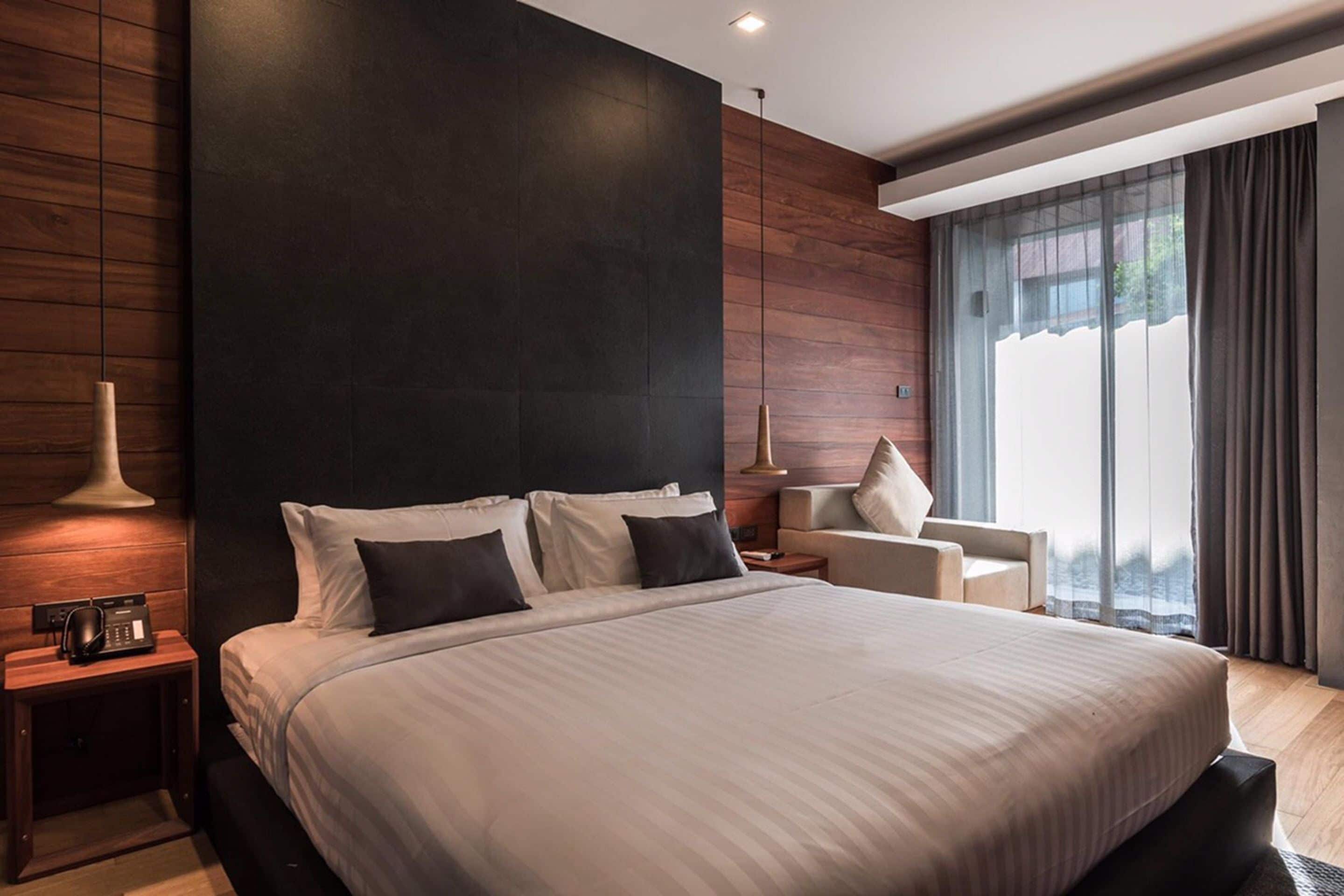 A modern hotel room with a large bed, a bedside table with a lamp, a chair, and a large window with curtains.
