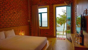 Quarto de hotel com parede de tijolos, uma cama grande, janela com vista para o mar e uma mesa com garrafa e copos.