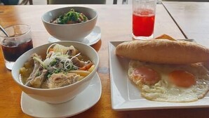 Uma mesa com uma refeição composta por sopa, sanduíche e bebida, com palmeiras ao fundo e vista para o mar.