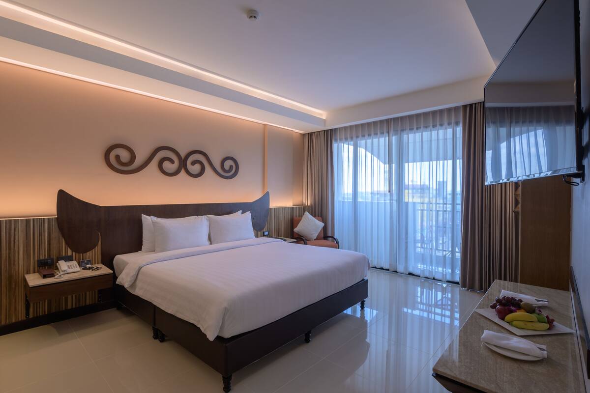 A modern hotel room with a large bed, a bedside table with a phone, a flat-screen TV, and a dining table with fruit.