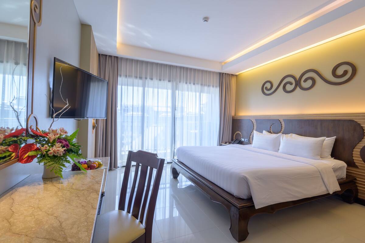 A hotel room with a large bed, a desk with a chair, a flat-screen TV mounted on the wall, and a large window with curtains.