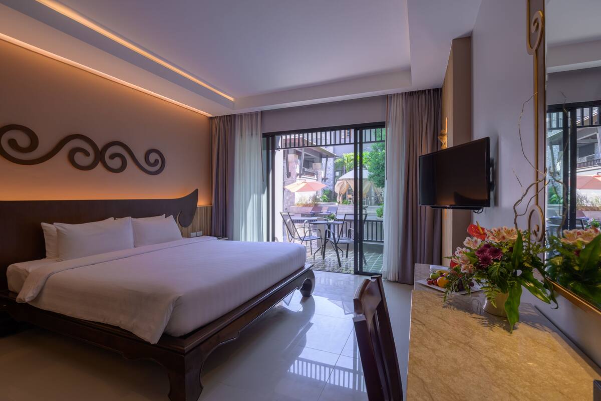 A hotel room with a large bed, a flat-screen TV mounted on the wall, a dining area with outdoor seating, and a balcony with a view of the outside.