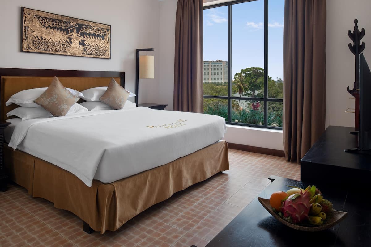 A hotel room with a large bed, a nightstand, a lamp, a window with curtains, and a view of buildings and trees.