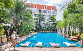 Una piscina con sillones de descanso, rodeada de plantas tropicales y un edificio con techo rojo al fondo.