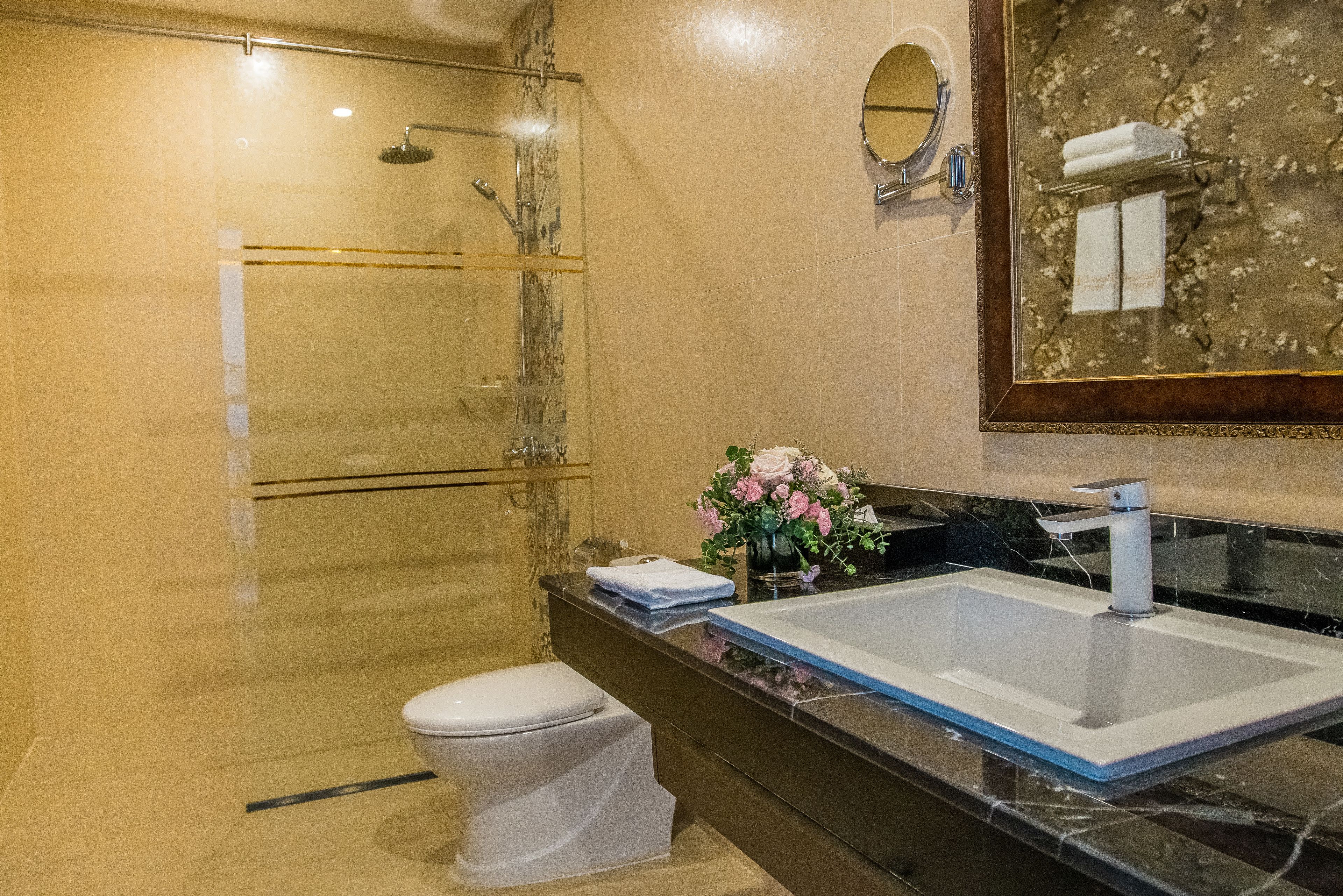 Een moderne badkamer met een douchecabine van glas, een wit toilet, een groot wastafelmeubel met een marmeren blad en een spiegel met een bloemmotief.