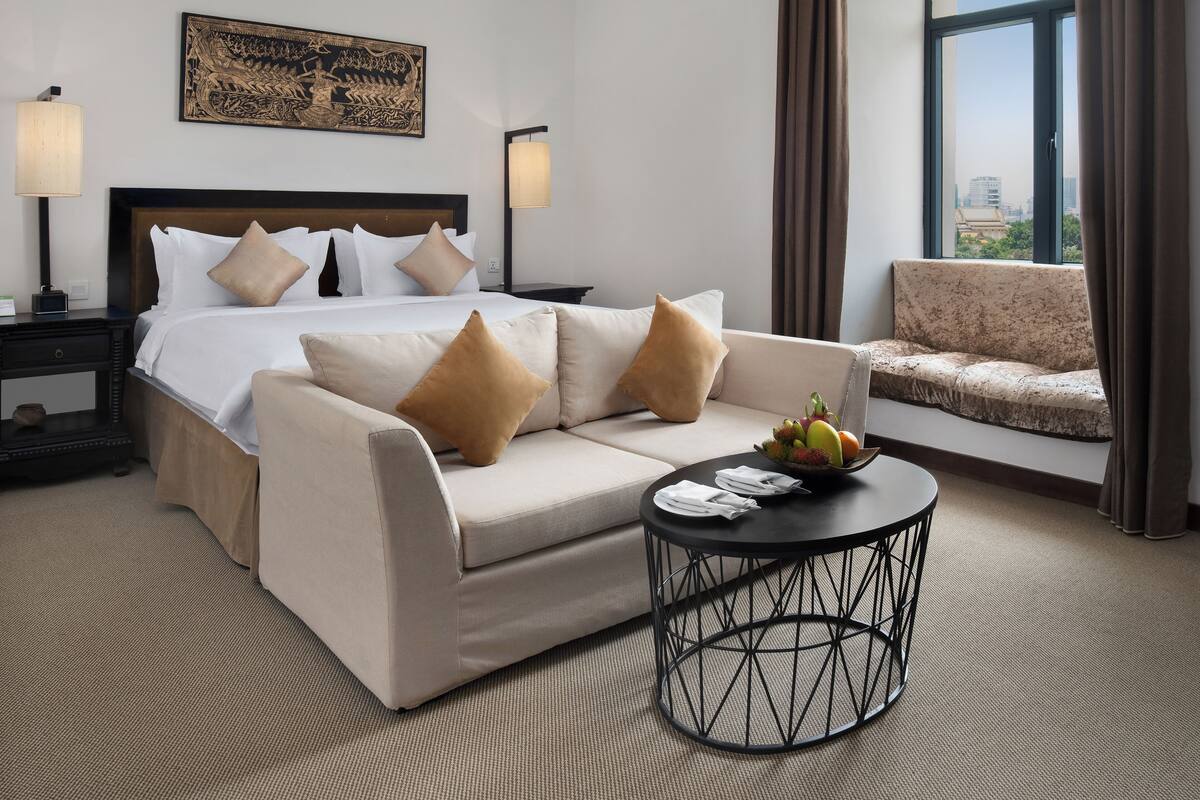 A hotel room with a bed, a sofa, a round coffee table, and a view of the city.