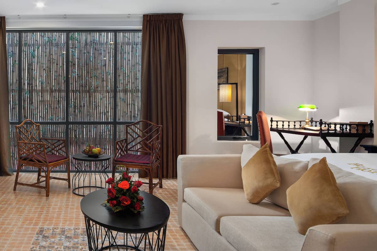 A hotel room with a sofa, two chairs, a small table with a vase of flowers, a bed with a white bedspread, and a view of a balcony with a bamboo screen.