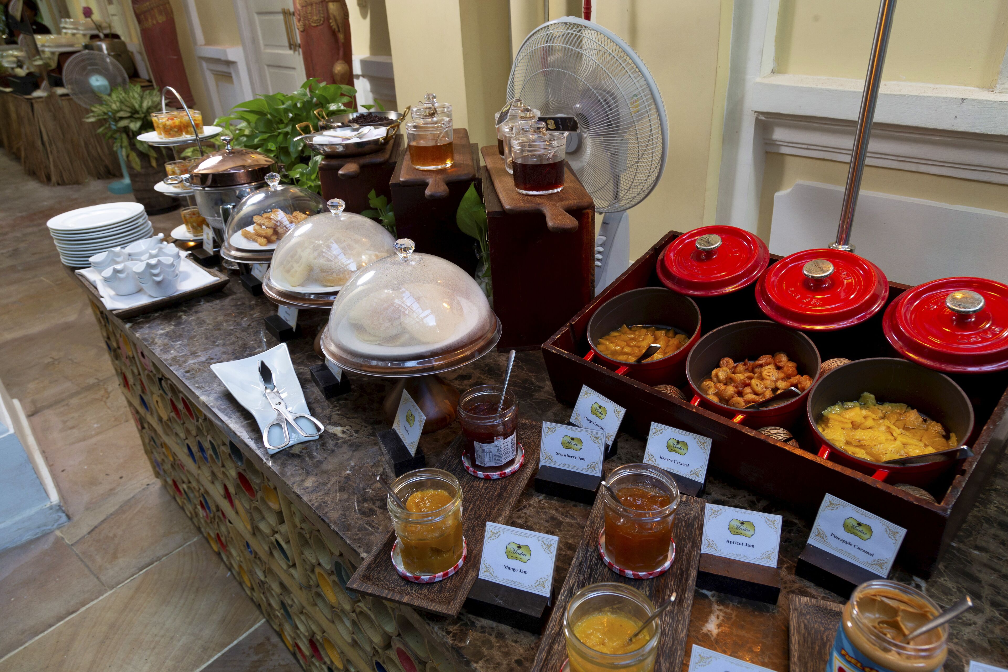 A buffet with various food items and condiments, including jars of jam and honey, covered dishes, and a fan for cooling.