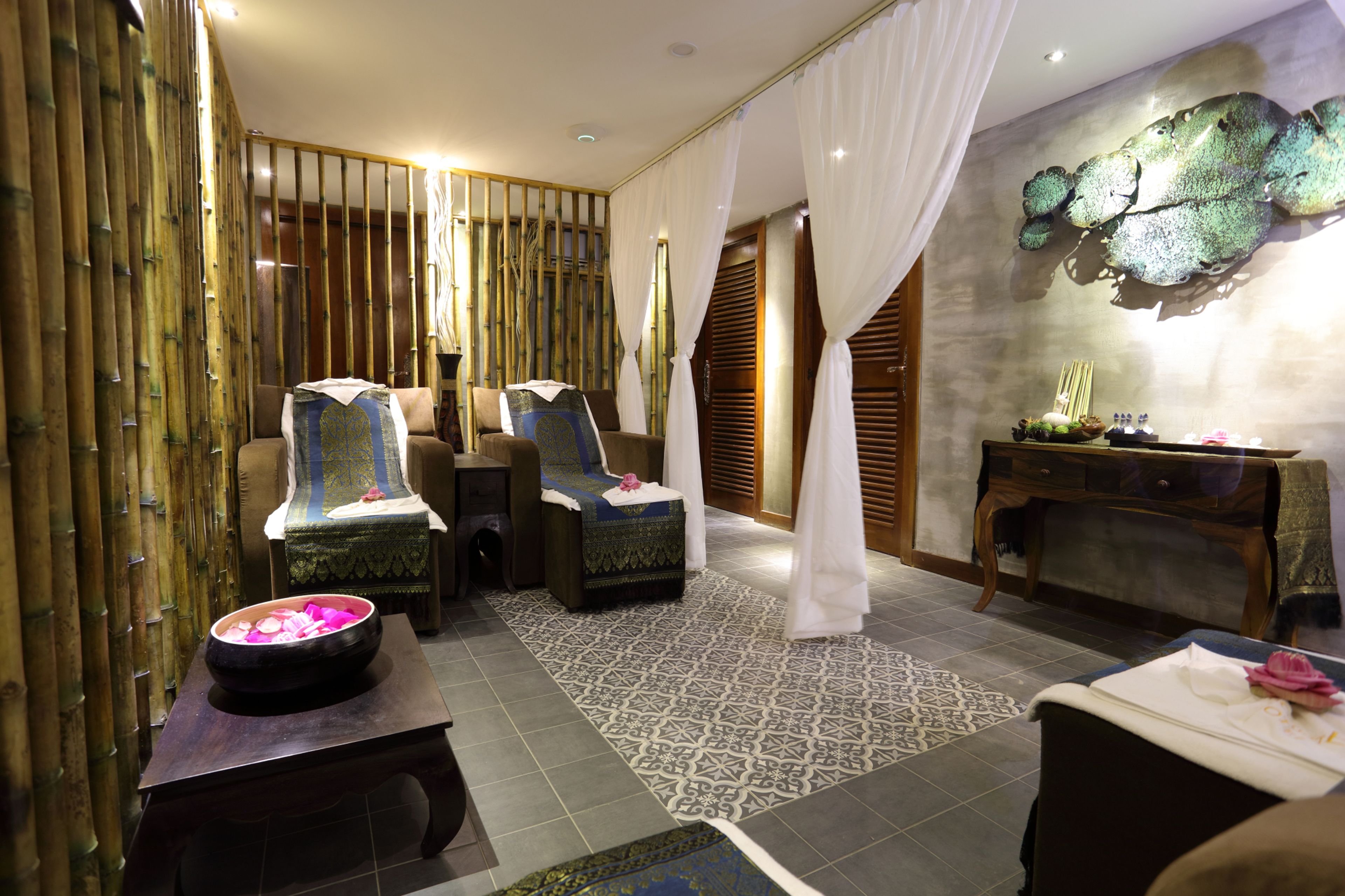 A spa room with two massage chairs, a small table with a bowl of rose petals, a wooden console table, and a wall-mounted sculpture.
