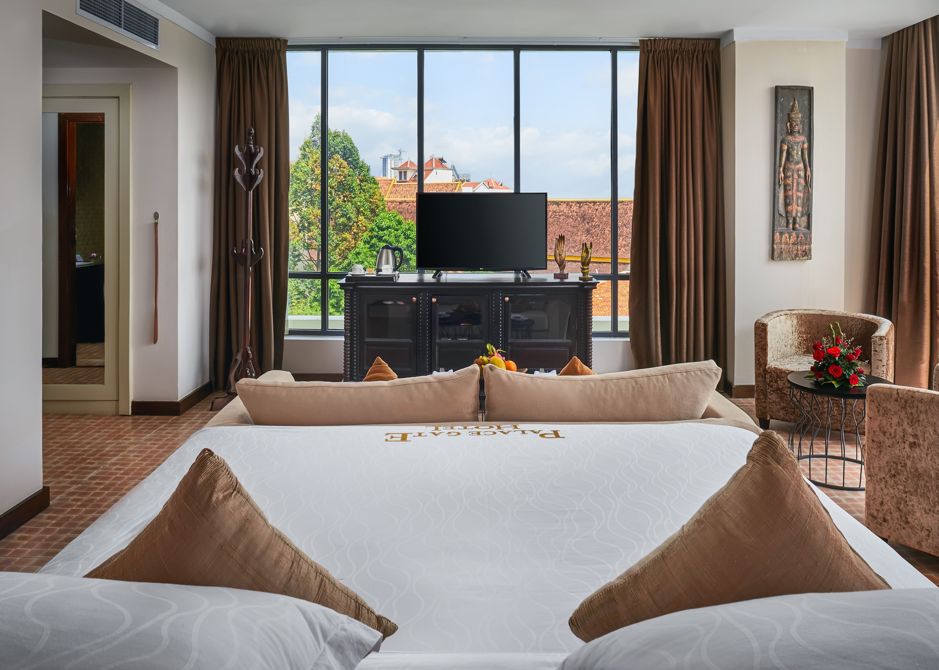 A hotel room with a large bed, a television on a cabinet, a chair with a flower arrangement, and a view of buildings and trees through the window.