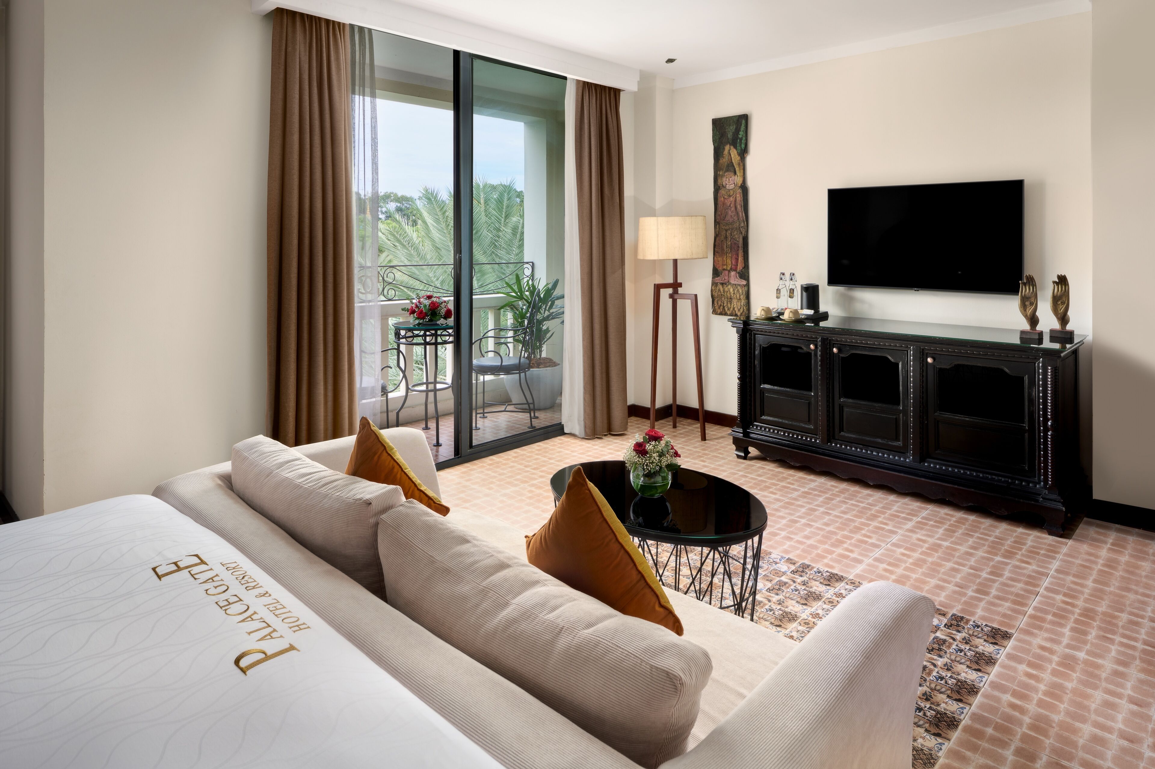 A hotel room with a large bed, a sofa, a small table with a vase, a TV, a cabinet, and a balcony with a table and chairs.