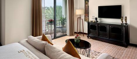 A hotel room with a large bed, a sofa, a small table with a vase, a TV, a cabinet, and a balcony with a table and chairs.