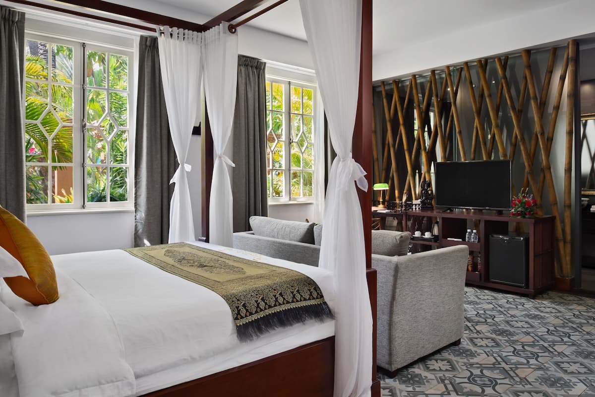 A hotel room with a bed, a television, a sofa, and a view of greenery through the windows.