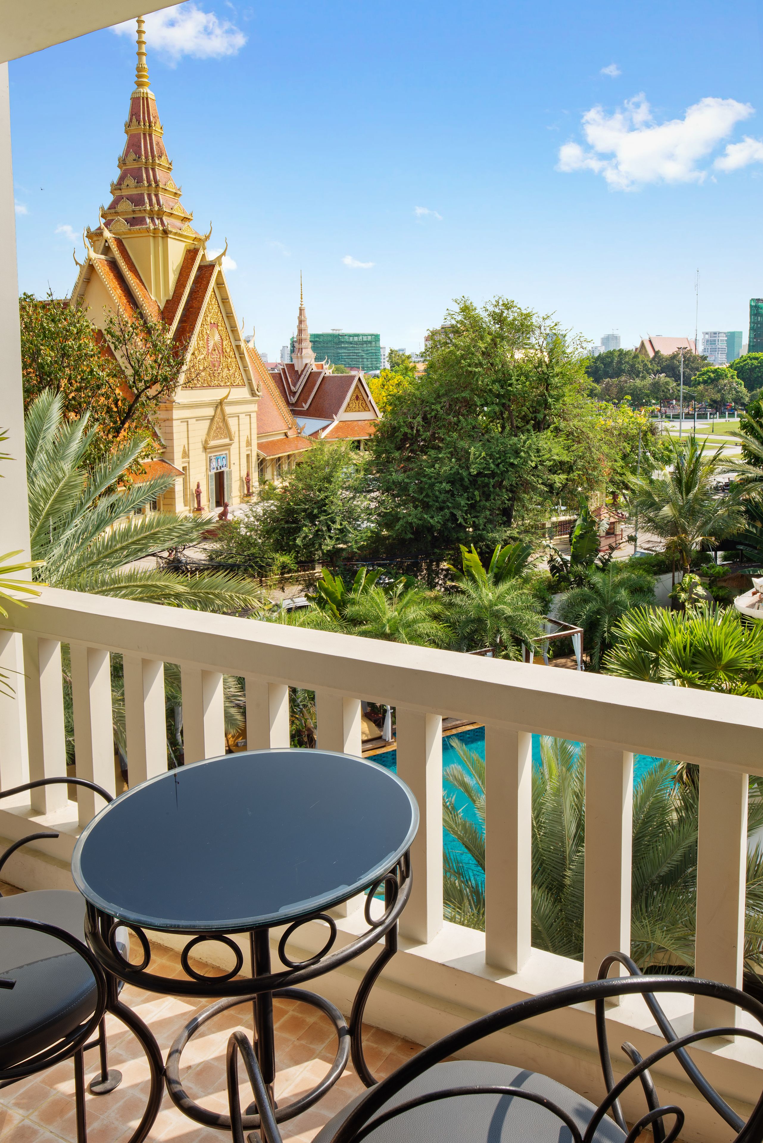 A balcony with a table and chairs, overlooking a temple with a pointed spire and a garden with palm trees.
