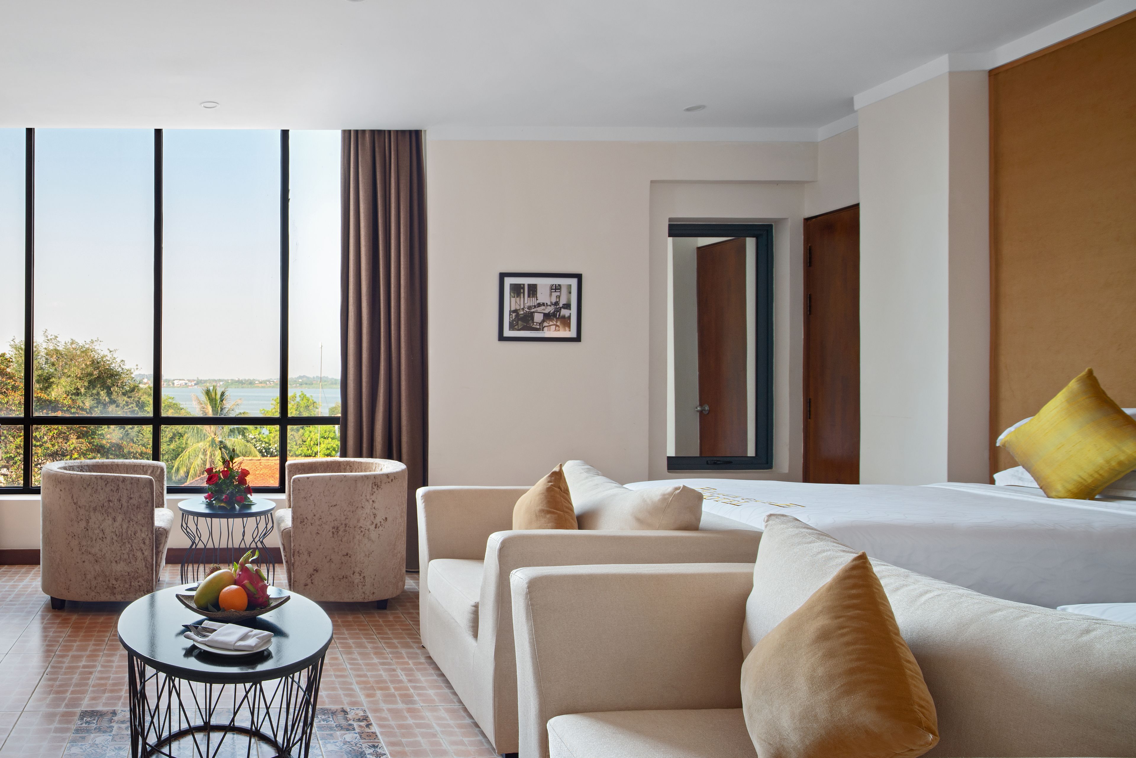 A hotel room with a large window, a bed, two sofas, a small table with fruit, and a picture on the wall.