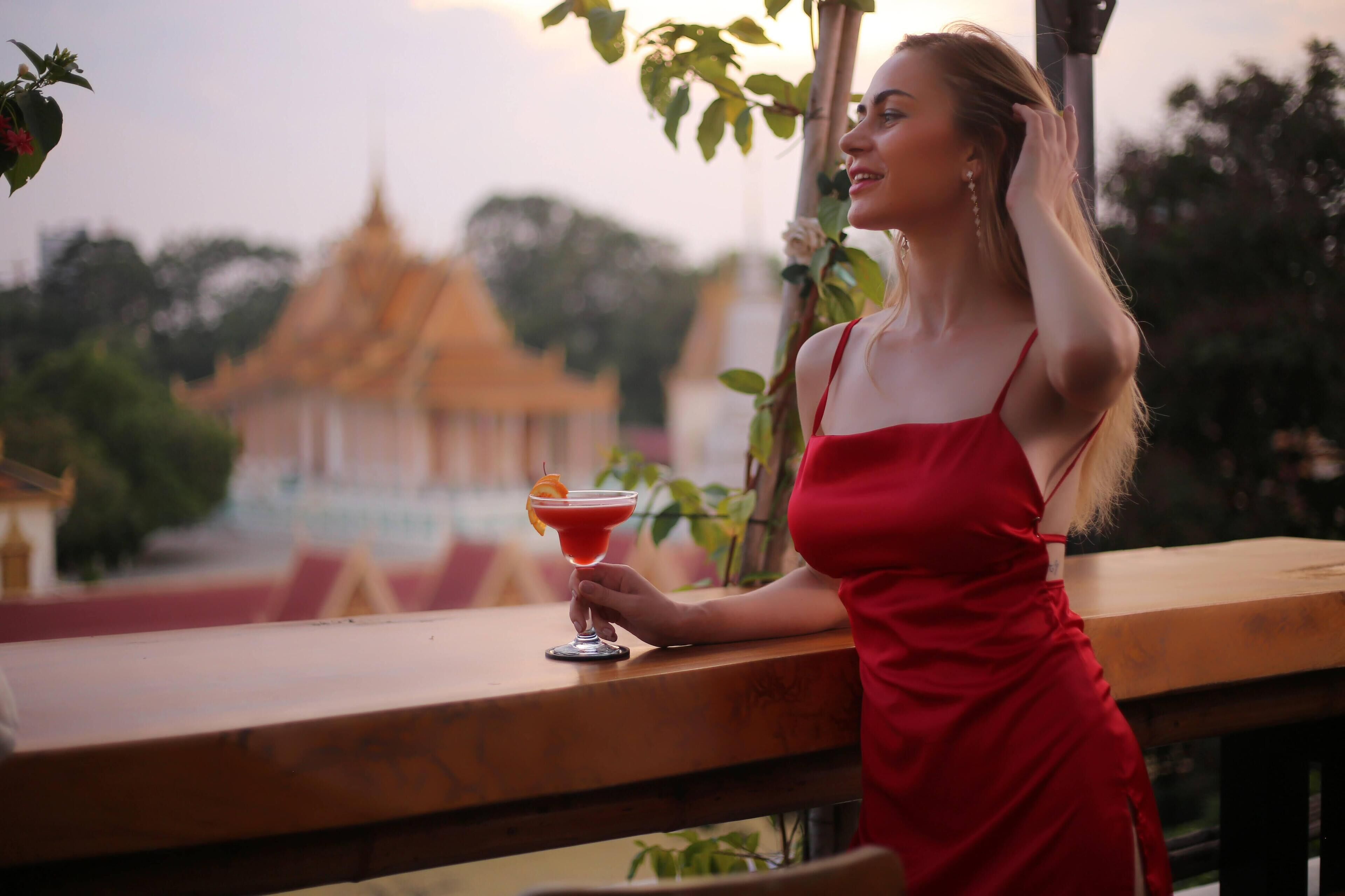 Una persona con un vestido rojo sosteniendo un cóctel en un balcón, con una vista de un templo al fondo.
