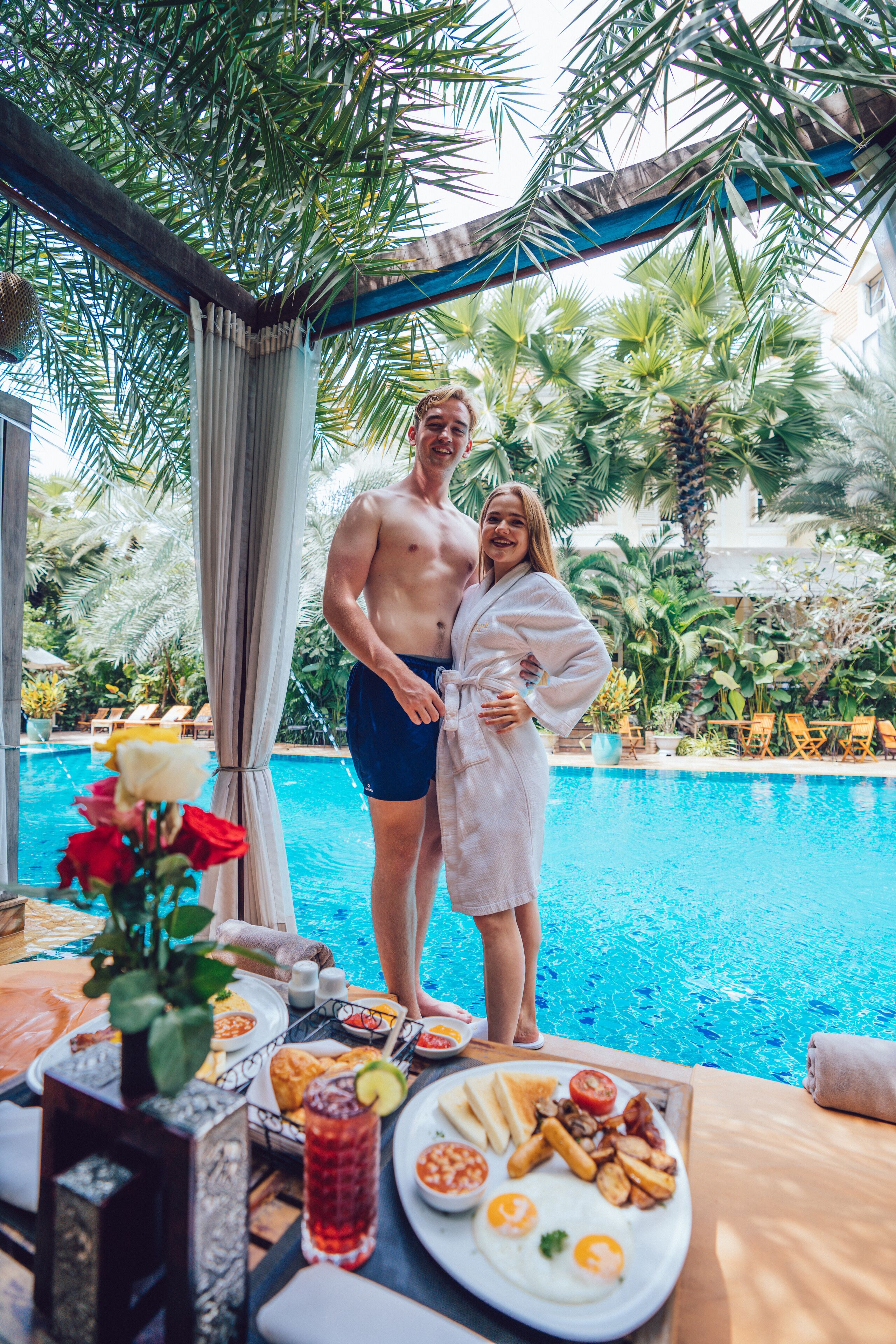A couple by a pool with a breakfast spread including eggs, toast, and fruit.