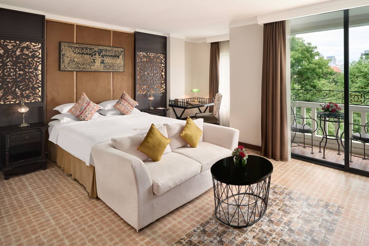 A hotel room with a large bed, a sofa, a small table, and a balcony with a view of trees and buildings.