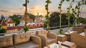 A rooftop terrace with wicker furniture, a view of a temple complex, and a sunset. - Palace Gate Hotel & Resort by EHM (Phnom Penh)