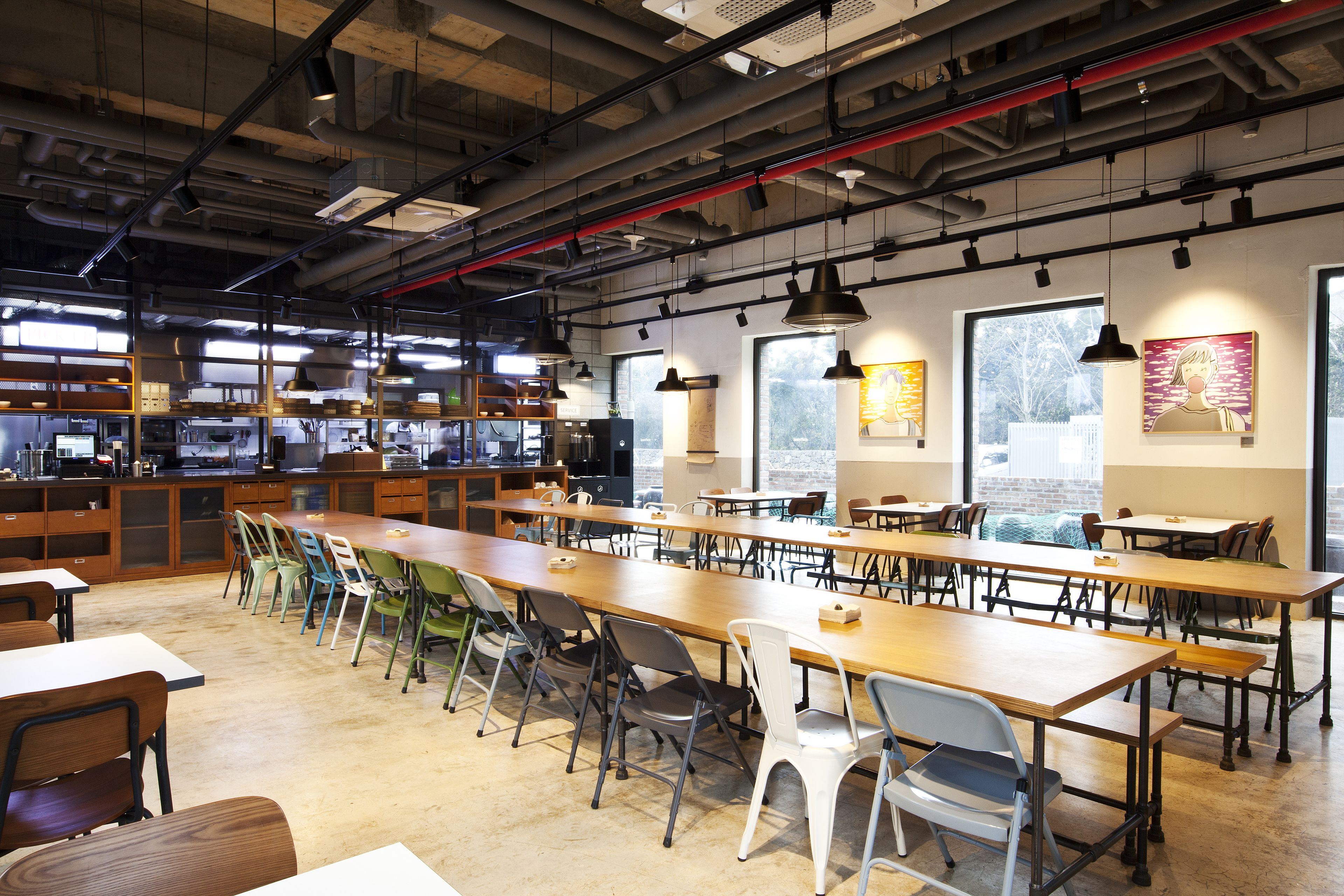 A spacious dining area with long tables and chairs, large windows, and industrial-style ceiling with exposed pipes.