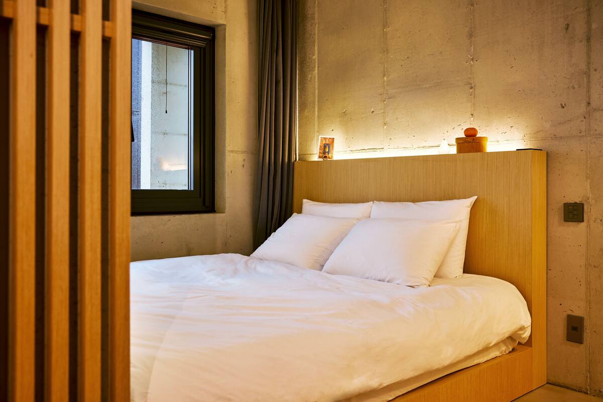 A modern bedroom with a wooden headboard, a large window, and a small bedside table with a lamp.